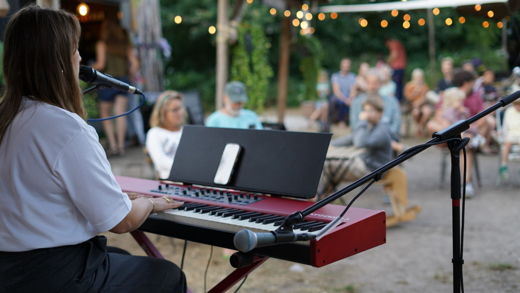 Nina speelt keyboard tijdens een buitenoptreden, gezien over haar schouder richting het publiek en de lichtjes
