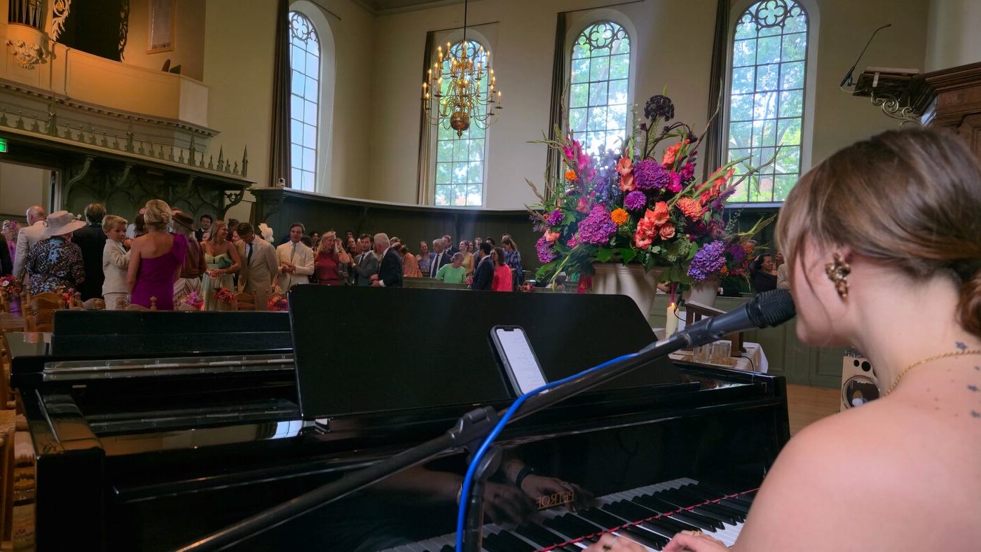 Nina speelt piano tijdens een kerkceremonie, gezien over haar schouder richting de gasten en een grote bloemenpracht