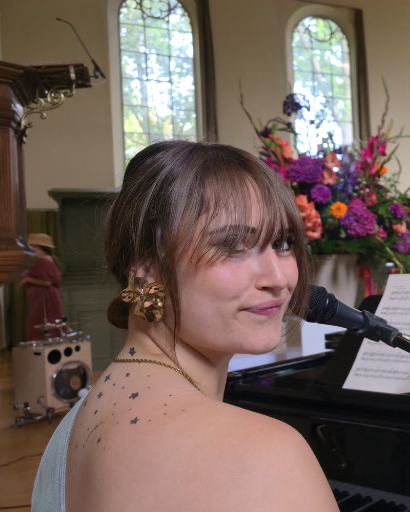 Nina kijkt om vanachter de piano tijdens een kerkceremonie, met een bloemstuk en de boogramen achter haar