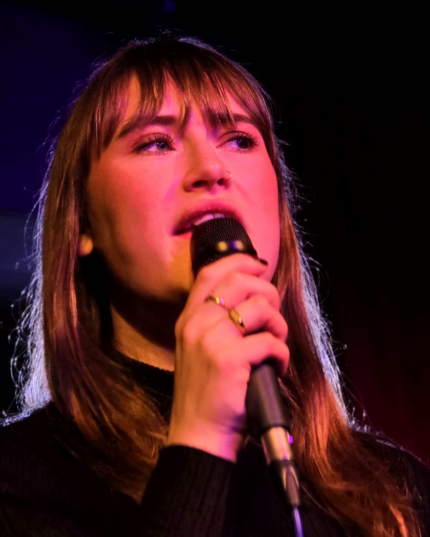 Nina zingt in een handmicrofoon tijdens een optreden, in rood podiumlicht