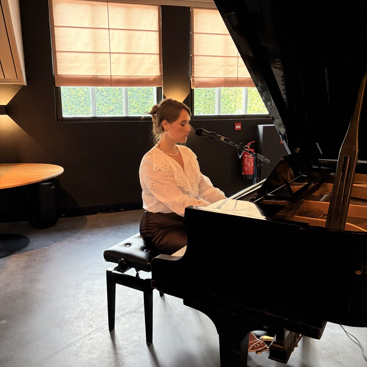 Nina speelt vleugel achter een microfoon in een zaal, met daglicht door de ramen